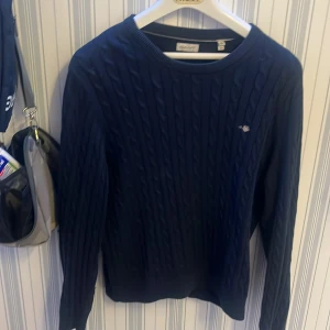 Mörkblå kabelstickad tröja från Gant - Säljer en stilren mörkblå kabelstickad tröja från Gant. Tröjan har en klassisk rund hals och långa ärmar, perfekt för kyligare dagar. Den är tillverkad i Turkiet och passar för en ungdom med storlek 170 cm.