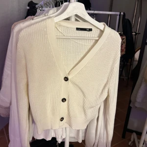 Vit stickad kofta från 157 - Säljer en stilren vit stickad kofta från 157. Koftan har en v-ringad design med knappar framtill och ribbade muddar vid ärmslut och nederkant. Perfekt för lager-på-lager under kyligare dagar.