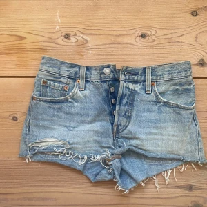 Blå jeansshorts från Levi's - Säljer ett par klassiska blå jeansshorts från Levi's med fransiga kanter och knappgylf. Perfekta för en avslappnad sommarlook.