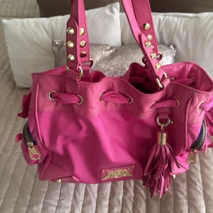 Rosa axelväska med nitar och tofs - Snygg rosa axelväska med coola nitar på handtagen och en dekorativ tofs. Väskan har dragkedjor och en rosett på sidan för extra stil. Perfekt för att ge din outfit en färgklick! Vintage Juicy Couture. Köpte den för många år sedan för 2700 kr men knappt använt den. 