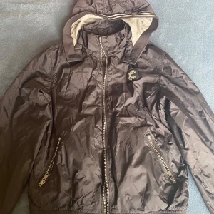 Svart vindjacka med huva - Säljer en svart vindjacka med huva och dragkedja framtill. Jackan har två fickor med dragkedjor och en ribbad kant vid ärmslut och nederkant. Perfekt för blåsiga dagar!