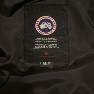 Svart dunväst från Canada Goose - Säljer en svart dunväst från Canada Goose med deras ikoniska logga på bröstet. Västen har en hög krage och knappar framtill. Perfekt för kyliga dagar.
