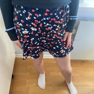 Blommig kortkjol från WE Fashion - Säljer en söt blommig kortkjol från WE Fashion. Kjolen har ett färgglatt mönster med blommor i rött, vitt och blått på en mörkblå bakgrund. Perfekt för en somrig look! 🌸 Den har också inbyggda byxor så du kan röra dig fritt❤️