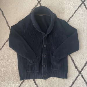 Mörkblå stickad kofta från Dustin - Säljer en snygg mörkblå stickad kofta från Dustin. Koftan har långa ärmar och knappar framtill, perfekt för kyliga dagar. Den har en stilren design med ribbade detaljer vid ärmslut och nederkant.