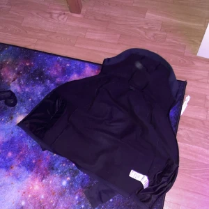 Svart/Mörk blå tröja från Peak Performance - Säljer en svart/mörk blå tröja från Peak Performance med dragkedja och huva. Perfekt för en sportig look eller som ett extra lager under kyliga dagar. Tröjan har en stilren design med logotypdetaljer.