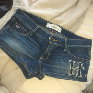 Blå jeansshorts från Hollister - Säljer ett par snygga blå jeansshorts från Hollister med ett stort 'H' broderat på framsidan. Shortsen har en klassisk femficksdesign och är tillverkade i stretchigt denim för extra komfort. Perfekta för en avslappnad sommarlook!