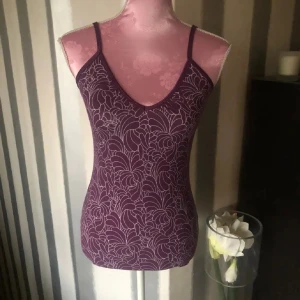 Lila mönstrad linnetopp - Säljer en snygg lila linnetopp med blommigt mönster. Toppen har smala axelband och en v-ringad halslinje. Perfekt för varma dagar eller som ett lager under en tröja. Med inbyggd bh