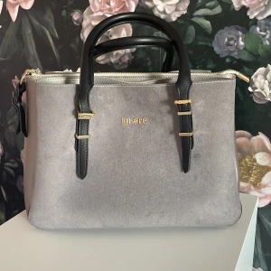 Grå handväska från BHPC i mocka-look - Snygg grå handväska från BHPC med svart handtag och gulddetaljer. Väskan har flera fack, dragkedja upptill och stilren design. Materialet har en mocka-liknande yta och väskan har justerbara spännen på handtagen. Perfekt för dig som gillar klassisk men modern stil.