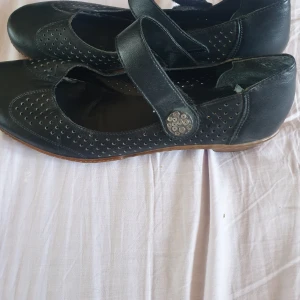 Svarta ballerinaskor från Rieker - Svarta ballerinaskor från Rieker med perforerat mönster och bred rem över foten som stängs med en dekorativ knapp. Skorna har rund tå och platt sula, tillverkade i skinn för en klassisk och bekväm look. Oanvända,stått I garderob pga jag inte kan använda de då jag använder fotinlägg för plattfot. 