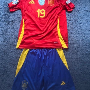 Spanien Lamine Yamal matchtröja Adidas - Säljer en officiell Spanien fotbollströja med Lamine Yamal och nummer 19. Tröjan är röd med gula detaljer och korta ärmar, tillverkad i lätt och ventilerande material. Adidas-logga och landslagsemblem framtill. Matchande blå shorts med gula inslag och landslagsemblem ingår.