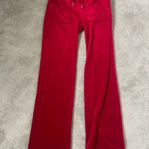 Röda velourbyxor Juicy Couture XXS - Röda velourbyxor från Juicy Couture med breda ben och stora fickor framtill. Byxorna har dragsko i midjan och klassisk logga broderad på ena fickan. Supermjuk känsla och ikonisk 2000-talsstil. OBS! Det finns en liten limfläck på framsidan av benet, men syns knappt.