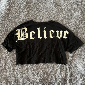 Svart croppad t-shirt från FB Sister - Cool svart croppad t-shirt från FB Sister i bomull. Stor text 'Believe' i vit gotisk stil på ryggen. T-shirten har korta ärmar och avslappnad passform, perfekt för en edgy streetstyle-look. Den har ett hål under höger arm (se bilden)
