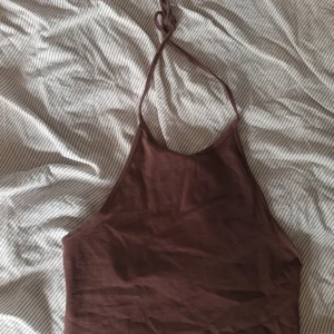 Brun halterneck linnetopp - Säljer en brun halterneck linnetopp med smala axelband som knyts i nacken. Toppen har en enkel och stilren design med hög halsringning och ärmlös passform. Perfekt för varma dagar och enkel att matcha med jeans eller kjol.