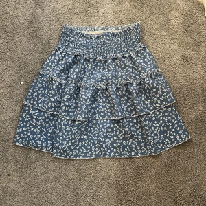 Blå blommig volangkjol - Supersöt blå kortkjol med vita små blommor och flera volanger. Kjolen har smock i midjan för extra bekväm passform och är perfekt för vår och sommar. Lätt och luftig känsla, passar till sneakers eller sandaler.