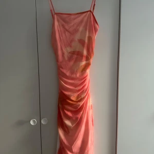 Blommig klänning med öppen rygg - Snygg klänning i aprkos och orange toner med blommigt mönster. Klänningen har smala axelband, draperad effekt och en öppen rygg med twist-detalj. Materialet är tunt och flowy, perfekt för varma dagar