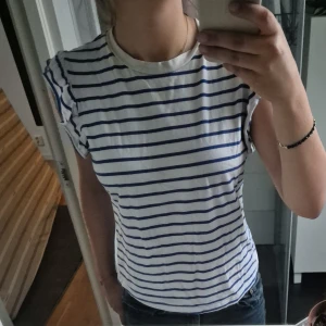 Randig vit och blå linnetopp - Säljer en fräsch vit linnetopp med blå horisontella ränder och volangdetaljer vid ärmhålen. Toppen har rund halsringning och är ärmlös, perfekt för en chill och stilren look. 