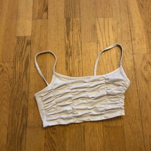 Beige croppad linnetopp med rynk - Snygg croppad linnetopp i beige med smala axelband och rynkade detaljer framtill. Perfekt för varma dagar och enkel att matcha med jeans eller kjol. Toppens material känns mjukt och följsamt mot huden.