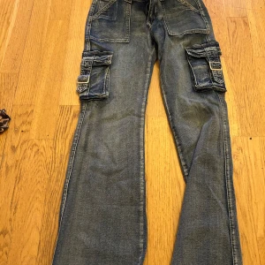 Blå cargojeans med fickor och spännen - Säljer ett par blå cargojeans med stora fickor på sidorna och coola spänndetaljer. Jeansen har en avslappnad passform och är perfekta för en streetwear-look. Materialet är klassisk denim och färgen är mellanblå.