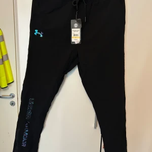 Svarta träningsbyxor Under Armour - Svarta träningsbyxor från Under Armour med snyggt holografiskt logotryck på benet och logga på höften. Byxorna har dragsko i midjan och är gjorda i ett lätt och svalt syntetmaterial, perfekt för träning. Regular fit och HeatGear-teknologi för att hålla dig torr.