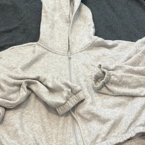 Ljusgrå hoodie med dragkedja, stl S - Mysig ljusgrå hoodie i mjuk bomullsblandning med dragkedja framtill och huva. Tröjan har långa ärmar med resår vid handlederna och en avslappnad passform. Perfekt för chill dagar eller när du vill vara extra bekväm.