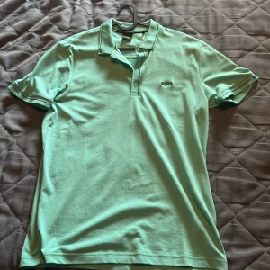 Mintgrön pikétröja från BOSS - Fräsch mintgrön pikétröja från BOSS i slim fit med klassisk krage och knappar framtill. Tröjan har diskret BOSS-logga på bröstet och snygga kontrastdetaljer vid krage och ärmslut. Tillverkad i mjuk bomull, perfekt för vår och sommar.