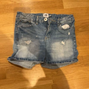 Blå jeansshorts från 157, storlek 160 - Snygga blå jeansshorts från 157 i storlek 160. De har slitna detaljer framtill, klassisk femficksmodell och uppvikta benslut. Perfekta för sommaren och har en normal passform. Gjorda i mjukt denimtyg med knapp och dragkedja framtill.
