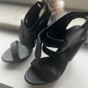 Svarta sandaler med klack från United Nude - Unika svarta sandaler från United Nude med hög klack och plattform. Skorna har breda remmar, öppen tå och cool futuristisk design. Tillverkade i skinn och syntet med både matta och blanka detaljer. Perfekta för dig som vill sticka ut med din stil.