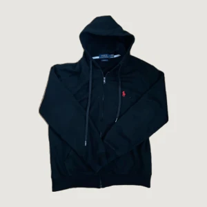 Svart hoodie från Polo Ralph Lauren - Svart hoodie från Polo Ralph Lauren med dragkedja och huva. Klassisk röd logga broderad på bröstet och snörning i huvan. Perfekt för en chill och stilren look. Tillverkad i mjuk bomull för extra komfort.