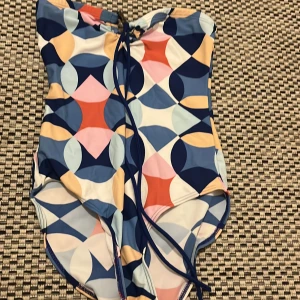 Färgglad baddräkt med snörning - Snygg baddräkt med grafiskt mönster i blått, rosa, rött, beige och vitt. Modellen har snörning framtill och är tillverkad i ett stretchigt syntetmaterial som passar perfekt för strand eller pool. Färgerna och mönstret ger en riktigt retro och lekfull vibe.