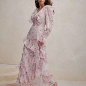 Ljusrosa blommig långklänning med volang - Romantisk långklänning i ljusrosa med blommigt mönster och volangdetaljer. Klänningen har v-ringning, långa ärmar och är tillverkad i ett lätt, flowy material som ger en drömmig känsla. Perfekt för sommarens alla tillfällen.