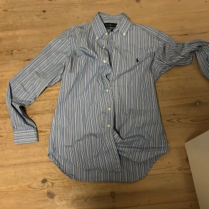 Blåvit randig skjorta från Ralph Lauren - Klassisk blå skjorta med vita och ljusblå ränder från Ralph Lauren. Skjortan har lång ärm, knappar framtill och button down-krage. Perfekt för sommaren