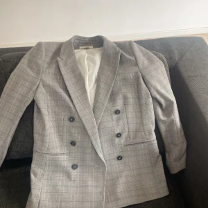 Grå rutig kavaj från H&M, stl 38 - Snygg grå kavaj från H&M i klassiskt rutig mönster. Dubbelknäppt med svarta knappar och två fickor framtill. Perfekt för en stilren look. Fodrad insida i vitt tyg och markerade axlar för en modern siluett.