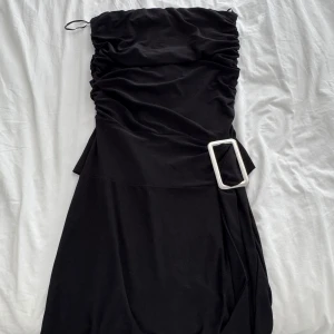 Svart kort klänning Sisters Point - Supersnygg svart kort klänning från Sisters Point med draperad överdel och stor vit spänndetalj i midjan. Axelbandslös modell som ger en cool och trendig look. Perfekt för dig som vill sticka ut med en enkel men stilren outfit.