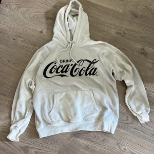 Vit Coca-Cola hoodie med tryck - Säljer en vit hoodie från Coca-Cola med stor svart logga framtill. Tröjan har huva med snörning och en klassisk känguruficka. Perfekt för dig som gillar streetwear och vill sticka ut med en ikonisk look.