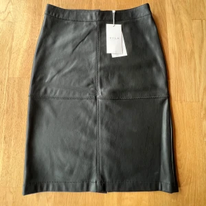 Svart skinnkjol från Vila - Snygg oanvänd svart skinnkjol från Vila med raka sömmar och dragkedja bak. Kjolen har en klassisk, figurnära passform och är tillverkad i ett mjukt skinnmaterial. 