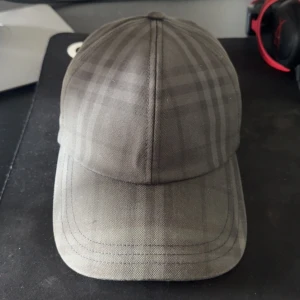 Rutig keps från Burberry i grått - Snygg keps från Burberry i grå toner med klassiskt rutigt mönster. Justerbar rem baktill med metallspänne och broderad Burberry-logga. Tillverkad i bomull, perfekt för dig som vill ha en stilren och exklusiv look. Använd cirka fem gånger. Köpt på NK gbg, kvitto och tags finns. Priset är ej hugget i sten.
