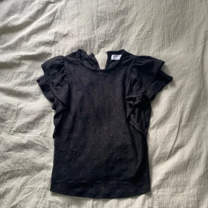 Svart blus från Lindex  - Supersöt svart blus från Lindex med diskret strukturmönster och voluminösa korta ärmar. Blusen har en liten v-ringning med knappdetalj i nacken och är tillverkad i ett mjukt tyg som känns skönt mot huden. Perfekt för dig som gillar stilrena detaljer.