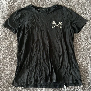 Svart t-shirt Keys To Freedom  - Svart t-shirt från The Black Label Collection i 100% bomull. Framsidan har tryck med två korsade nycklar och baksidan har större tryck med texten 'KEYS TO FREEDOM TBLC'. Klassisk passform och rund hals, perfekt för en chill look.