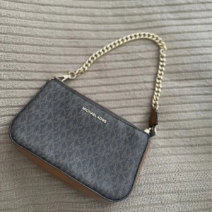 Michael Kors brun axelväska med kedja - Snygg axelväska från Michael Kors i mörkbrunt och beige med MK-mönster. Väskan har guldfärgad kedjerem, dragkedja upptill och detaljer i skinn. Perfekt storlek för det viktigaste och ger en lyxig känsla till din outfit.