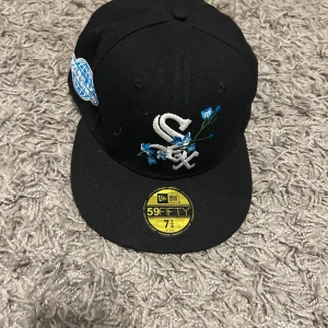 Svart New Era 59FIFTY keps White Sox - Snygg svart 59FIFTY keps från New Era med White Sox-logga framtill och broderade blå blommor. Sidopatch med World Series-märke i blått och vitt. Undersidan av skärmen är ljusblå med baseboll-detalj. Unik och perfekt för dig som vill sticka ut med stil. Tveka inte på att skriva vid minsta fundering.