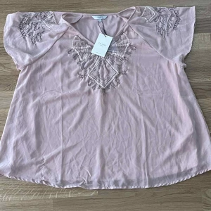 Ljusrosa blus med pärldetaljer Cellbes - Supersöt ljusrosa blus från Cellbes med v-ringning och korta, vida ärmar. Blusen har broderade detaljer och pärlor framtill samt på axlarna som ger en extra touch. Perfekt för dig som gillar feminina och unika plagg.