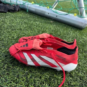 Adidas Predator röda fotbollsskor - Snygga Adidas Predator fotbollsskor i rött med vita och svarta detaljer. Skorna har snörning, låg profil och mönstrad sula för bästa grepp på planen. Ovandelen är i syntetmaterial och designen är sportig med tydliga Predator-loggor.