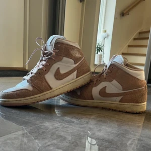 Nike Air Jordan 1 i beige mocka - Nike Air Jordan 1 sneakers i en snygg mix av beige mocka och vitt skinn. Klassisk high-top siluett med rund tå, platt sula och ikoniska vingar-loggan på sidan. Perfekta för dig som vill sticka ut med stilrena och trendiga sneakers.