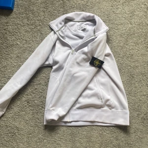 Vit fleecejacka från Stone Island M - Vit fleecejacka från Stone Island med klassisk logga på ärmen. Jackan har dragkedja framtill, två fickor med dragkedja och hög krage. Perfekt för dig som gillar stilren och clean look. Mjuk insida i fleece för extra komfort.