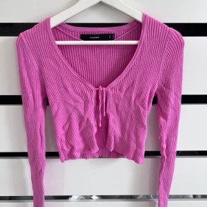 Rosa ribbad croppad topp Vero Moda - Superfin rosa croppad topp från Vero Moda med långa ärmar och snygg ribbad struktur. Toppen har v-ringning och en söt knytning framtill som ger en trendig vibe. Perfekt att styla med jeans eller kjol för en cool look.