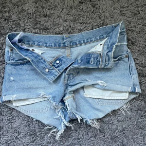 Levi's ljusblå jeansshorts med fransar - Ljusblå jeansshorts från Levi's med fransiga kanter och slitna detaljer. Klassisk femficksmodell med knappar i gylfen och Levi's läderpatch bak. Perfekta för en avslappnad stil under varma dagar. Är i storlek xs. 