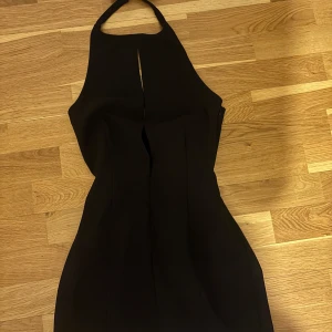 Svart halterneckklänning med rosett - Snygg svart kortklänning med halterneck och en dekorativ rosett med pärldetaljer i nacken. Klänningen har en stilren passform och en liten slits framtill som ger en cool touch. Perfekt för dig som gillar minimalistisk men ändå unik stil.