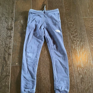 Blå mjukisbyxor från The North Face - Blå mjukisbyxor från The North Face med vit logga på låret. Byxorna har resår och snörning i midjan, ribbade muddar vid bensluten och är tillverkade i mjukt material med fleece på insidan. Perfekta för chill eller träning.