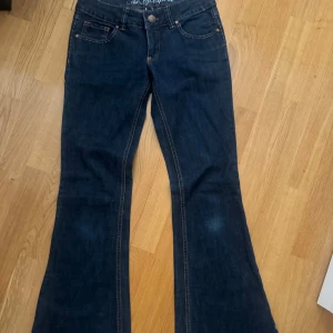 Mörkblå bootcut jeans från EDC - Snygga mörkblå jeans från EDC med bootcut passform och klassiska kontrastsömmar. Bakfickorna har broderade detaljer och ett läderpatch med logga i midjan. Perfekta för dig som gillar en retro vibe och vill ha ett par jeans med lite extra sväng i benen. Storlek 26/32