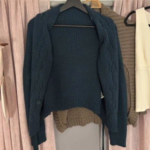Mörkgrön stickad kofta från Intimissimi - Mörkblå stickad kofta från Intimissimi med snyggt mönstrade partier längs framstyckena. Koftan har lång ärm, ribbade muddar och en knappdetalj och är knytbar vid ärmslutet. Perfekt för lager-på-lager och chill höst- eller vinterstil. I strlk s inte kommit till användning därav säljs den